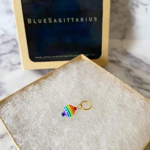 May include: A small, colorful charm with a rainbow design and a gold-colored ring. The charm is displayed in a small, open box with white padding. The box is next to a larger box with the text "BLUESAGITTARIUS".