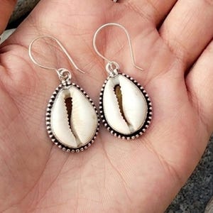 Puede incluir: Un par de pendientes colgantes plateados con colgantes en forma de lágrima. Cada colgante presenta una concha de cauri blanca con un interior marrón, enmarcada por un borde plateado con cuentas. Fondo de piel.