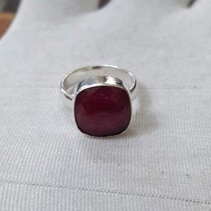 Könnte beinhalten: Ein Silberring mit einem großen, quadratisch geschliffenen, tiefroten Edelstein. Der Edelstein ist der Mittelpunkt, gefasst in einem schlichten, polierten Silberband. Der Ring wird auf einer weißen, strukturierten Oberfläche präsentiert.