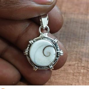 May include: A silver pendant with a white, spiral-patterned center stone. The pendant has a circular design with ornate silver detailing around the stone and a small loop at the top for a chain.
