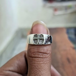 Pode incluir: Um anel de prata com um design de cruz. O anel é largo e tem um acabamento polido. A cruz está centrada no anel e tem uma aparência texturizada. O anel é mostrado no dedo de uma pessoa.