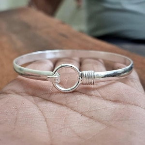 May include: A silver-colored bangle bracelet with a circular clasp and a wrapped section near the clasp. The bracelet is round and appears to be made of metal. The clasp is a simple hook and loop design.