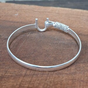 K&ouml;nnte beinhalten: Ein silbernes Armband mit Hakenverschluss und geflochtenem Detail. Das kreisf&ouml;rmige Armband liegt auf einer Holzoberfl&auml;che. Der Hakenverschluss befindet sich an einem Ende, das geflochtene Detail am anderen.
