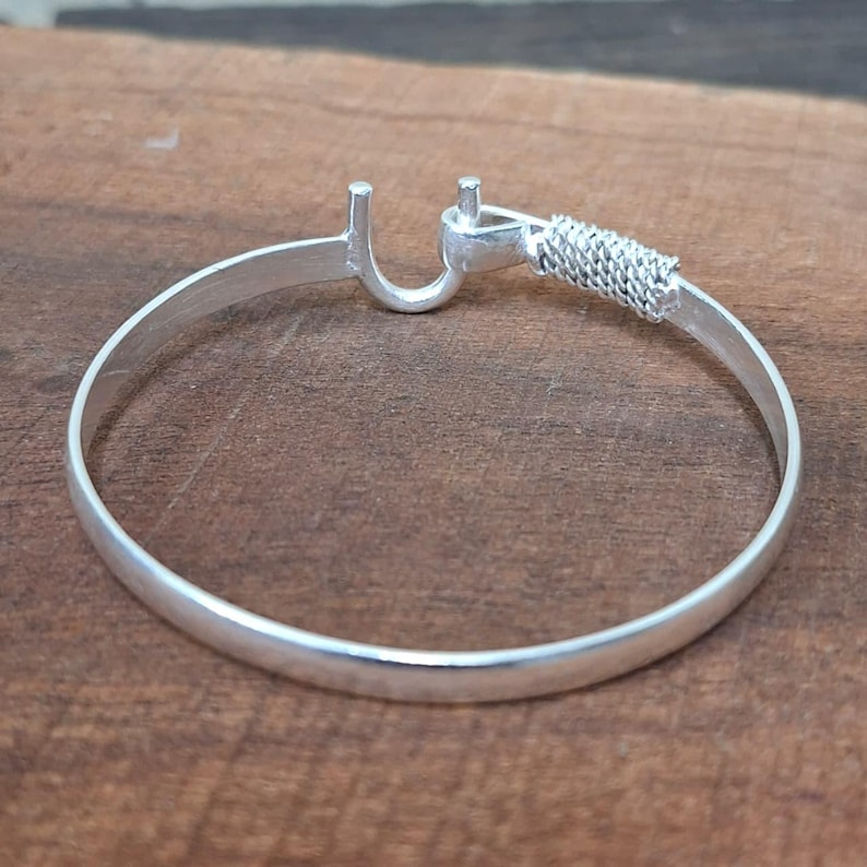 K&ouml;nnte beinhalten: Ein silbernes Armband mit Hakenverschluss und geflochtenem Detail. Das kreisf&ouml;rmige Armband liegt auf einer Holzoberfl&auml;che. Der Hakenverschluss befindet sich an einem Ende, das geflochtene Detail am anderen.