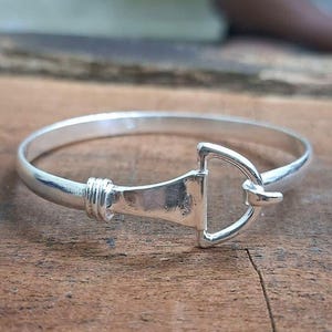 May include: A silver-colored bangle bracelet with a unique stirrup-shaped clasp. The bracelet is round and has a smooth, polished finish. The clasp is a prominent feature, adding a distinctive touch to the jewelry.