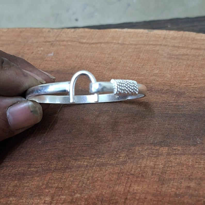 K&ouml;nnte beinhalten: Ein silberfarbenes Armband mit einem einzigartigen Design. Das Armband hat ein gebogenes, bogenf&ouml;rmiges Element und ein strukturiertes, zylindrisches Ende. Das Metall hat eine polierte Oberfl&auml;che, die das Licht reflektiert. Das Armband wird vor einem Holzuntergrund gezeigt.