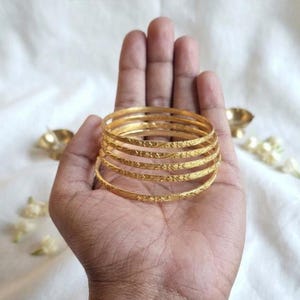 May include: A set of five gold-coloured metal bangles featuring a textured, etched surface pattern. The circular bracelets are stacked together, displaying a consistent metallic finish and intricate detailing along the outer circumference of each band.