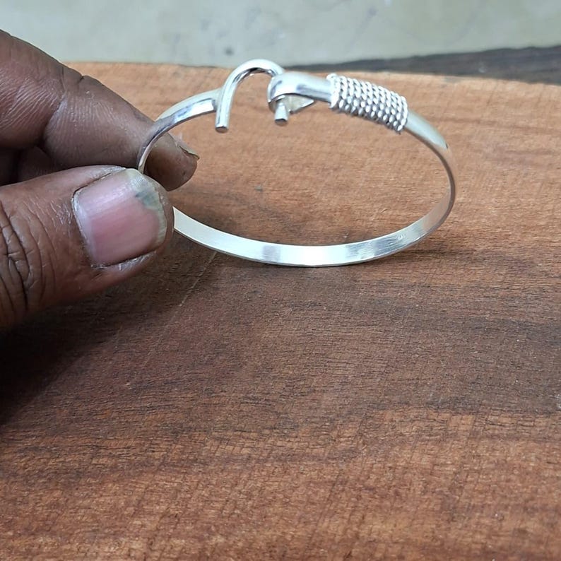 K&ouml;nnte beinhalten: Ein silberfarbenes Armband mit einem einzigartigen Haken- und Knotendesign. Das Armband hat eine gewebte Textur an einem Ende und ein schlichtes, elegantes Band. Der Hakenverschluss verleiht diesem Schmuckst&uuml;ck eine besondere Note.