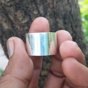 May include: A wide, silver-colored ring with a smooth, polished surface. The ring is held between two fingers, showcasing its simple, minimalist design. The ring's width appears to be approximately 0.5 inches.