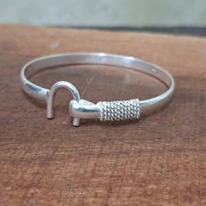 Puede incluir: Una pulsera plateada con cierre de gancho. La pulsera presenta un diseño sencillo con un cierre de gancho y un detalle de alambre tejido cerca del cierre. La pulsera se muestra sobre una superficie de madera.