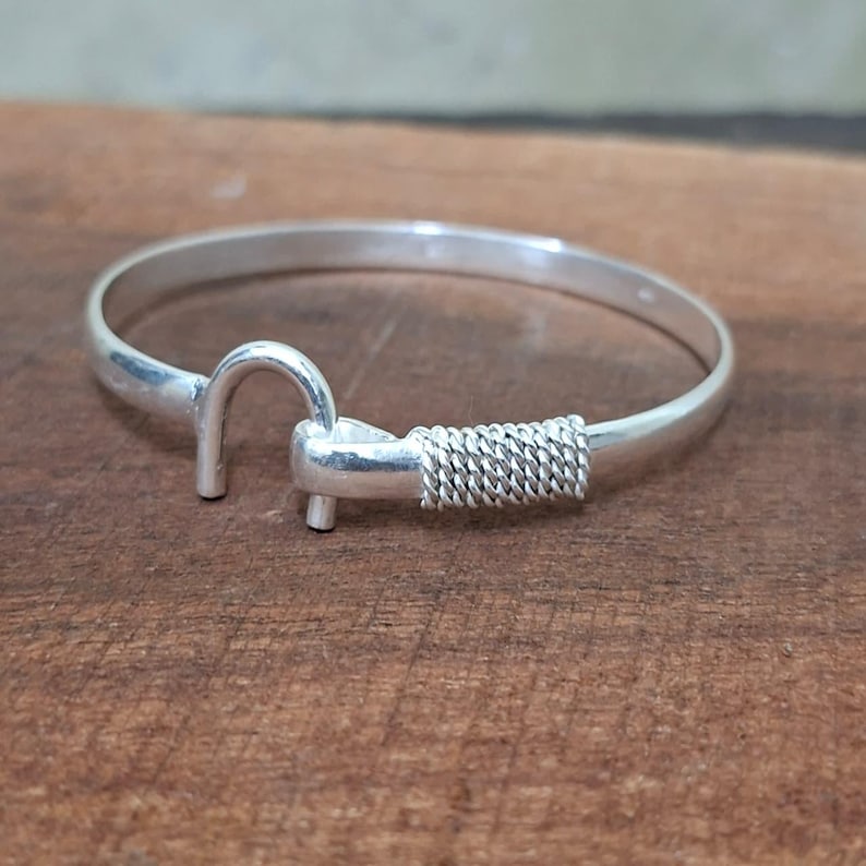 K&ouml;nnte beinhalten: Ein silberfarbenes Armband mit Hakenverschluss. Das Armband hat ein schlichtes Design mit einem Hakenverschluss und einem geflochtenen Drahtdetail in der N&auml;he des Verschlusses. Das Armband wird auf einer Holzoberfl&auml;che pr&auml;sentiert.