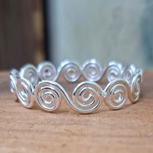 May include: A silver ring with a continuous swirl design. The ring features a series of connected spiral shapes, creating a wave-like pattern. The ring is made of a shiny, reflective metal, and is set against a blurred background.