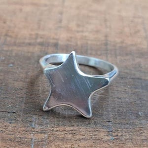 Puede incluir: Un anillo plateado con un diseño de estrella de cinco puntas. La estrella tiene una superficie lisa y pulida. La banda del anillo es simple y complementa la forma de la estrella. El anillo está sobre un fondo de madera.