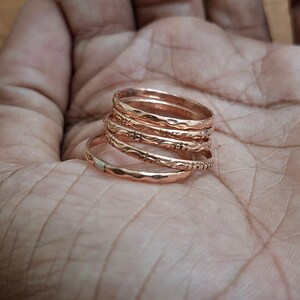 Könnte beinhalten: Ein Stapel von sechs roségoldenen Ringen mit strukturierten und gemusterten Designs. Die Ringe sind übereinander gestapelt und zeigen die verschiedenen Texturen und Muster. Die Ringe werden auf einem hautfarbenen Hintergrund präsentiert.