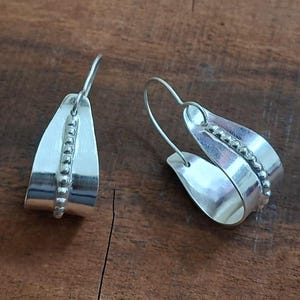 May include: A pair of silver earrings with a curved, layered design. Each earring features a row of small, round beads down the center. The earrings are displayed on a dark brown wooden surface.