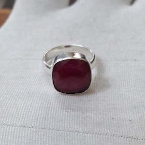 May include: A silver ring featuring a large, cushion-cut, deep red gemstone set in a polished metal band. The stone has a faceted surface that reflects light, and the ring is constructed with a smooth, rounded band that encircles the finger.