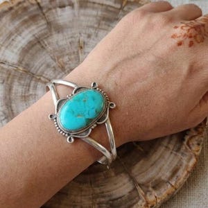 May include: A silver cuff bracelet featuring a large, oval-shaped turquoise stone set in a decorative metal frame. The stone displays a vibrant blue-green colour with natural veining. The metal band has a polished finish with intricate detailing.