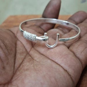 K&ouml;nnte beinhalten: Ein silberfarbenes Armreif-Armband mit Hakenverschluss. Das Armband hat ein gewebtes Design an einem Ende und einen Hakenverschluss am anderen. Ein schlichtes, elegantes Schmuckst&uuml;ck.