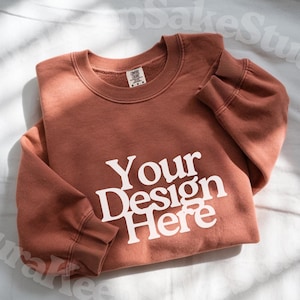 Pode incluir: Uma camisola de cor terracota com o texto branco "Your Design Here". A camisola de gola redonda est&aacute; dobrada sobre uma superf&iacute;cie branca. A imagem tamb&eacute;m inclui os log&oacute;tipos Canva, Photoshop e Procreate.