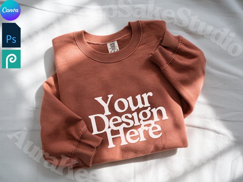 Pode incluir: Uma camisola de cor terracota com o texto branco "Your Design Here". A camisola de gola redonda est&aacute; dobrada sobre uma superf&iacute;cie branca. A imagem tamb&eacute;m inclui os log&oacute;tipos Canva, Photoshop e Procreate.