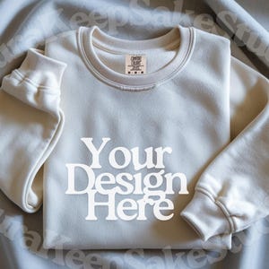 Pode incluir: Camisola creme com o texto "Your Design Here" em letras brancas. A camisola tem gola redonda e punhos canelados. Uma pequena etiqueta &eacute; vis&iacute;vel perto da gola. A camisola est&aacute; dobrada sobre um tecido cinza claro.