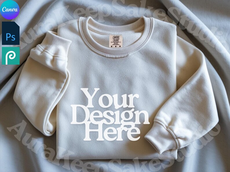 Pode incluir: Camisola creme com o texto "Your Design Here" em letras brancas. A camisola tem gola redonda e punhos canelados. Uma pequena etiqueta &eacute; vis&iacute;vel perto da gola. A camisola est&aacute; dobrada sobre um tecido cinza claro.
