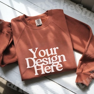 Pode incluir: Um moletom cor de terracota com o texto "Your Design Here" em branco. O moletom est&aacute; dobrado cuidadosamente sobre uma superf&iacute;cie de madeira branca. A imagem tamb&eacute;m inclui os logotipos Canva, Photoshop e Procreate.