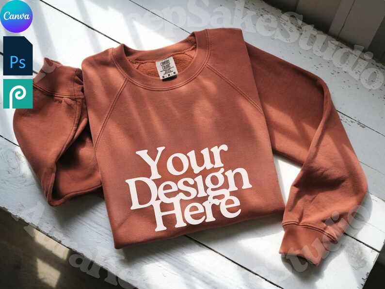 Pode incluir: Um moletom cor de terracota com o texto "Your Design Here" em branco. O moletom est&aacute; dobrado cuidadosamente sobre uma superf&iacute;cie de madeira branca. A imagem tamb&eacute;m inclui os logotipos Canva, Photoshop e Procreate.
