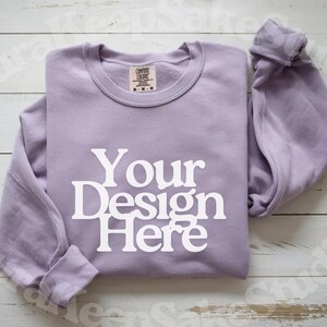 Pode incluir: Um moletom lavanda com gola redonda com o texto "Your Design Here" em branco. O moletom est&aacute; dobrado sobre uma superf&iacute;cie de madeira clara. A etiqueta da marca Comfort Colors &eacute; vis&iacute;vel na gola.