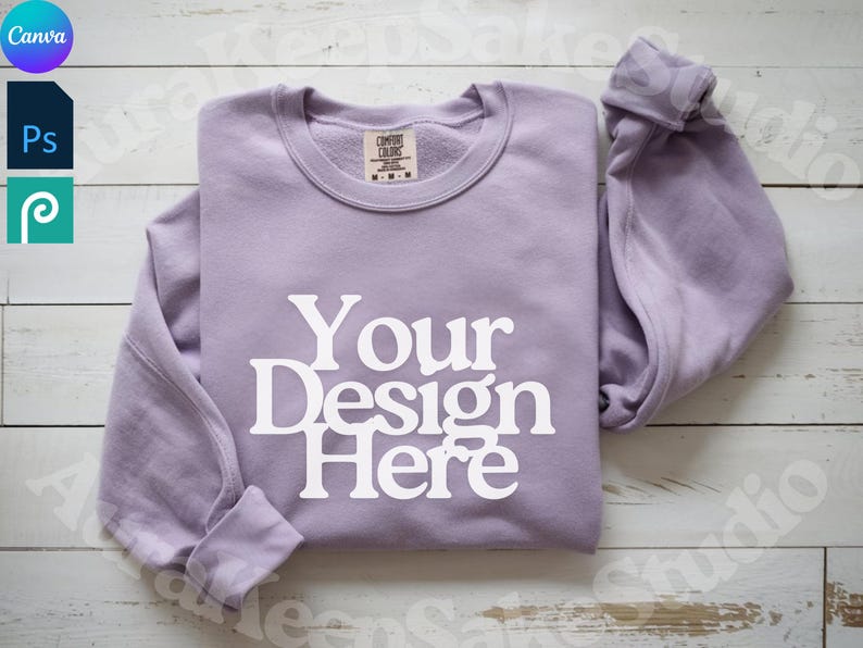 Pode incluir: Um moletom lavanda com gola redonda com o texto "Your Design Here" em branco. O moletom est&aacute; dobrado sobre uma superf&iacute;cie de madeira clara. A etiqueta da marca Comfort Colors &eacute; vis&iacute;vel na gola.