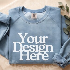 Pode incluir: Um moletom azul claro com gola redonda e o texto branco "Your Design Here". O moletom tem um visual desbotado e uma etiqueta "Comfort Colors". Um ramo de vegeta&ccedil;&atilde;o repousa ao lado.