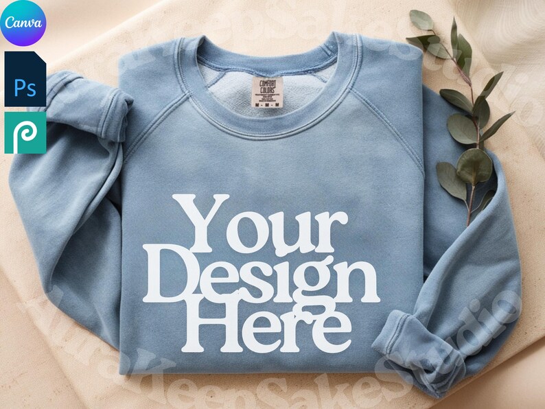 Pode incluir: Um moletom azul claro com gola redonda e o texto branco "Your Design Here". O moletom tem um visual desbotado e uma etiqueta "Comfort Colors". Um ramo de vegeta&ccedil;&atilde;o repousa ao lado.