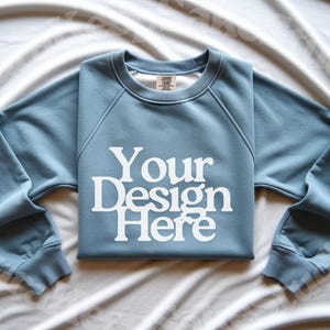 Pode incluir: Um moletom azul claro com o texto branco "Your Design Here". O moletom est&aacute; dobrado sobre uma superf&iacute;cie branca. O design est&aacute; centrado na frente. As mangas s&atilde;o longas e o decote &eacute; redondo.