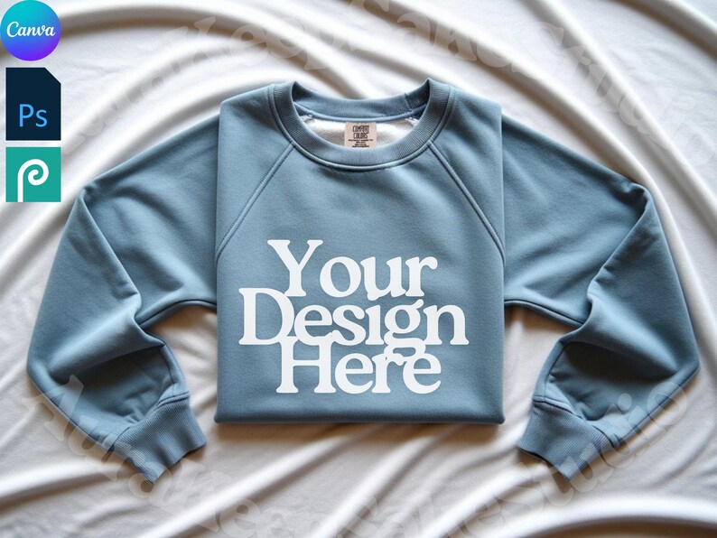 Pode incluir: Um moletom azul claro com o texto branco "Your Design Here". O moletom est&aacute; dobrado sobre uma superf&iacute;cie branca. O design est&aacute; centrado na frente. As mangas s&atilde;o longas e o decote &eacute; redondo.