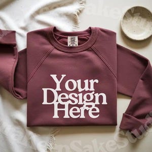 Pode incluir: Uma camisola de gola redonda bord&ocirc; com o texto "Your Design Here" em branco. A camisola &eacute; exibida em uma superf&iacute;cie branca com uma pequena tigela branca. As mangas est&atilde;o dobradas. Os logotipos Canva, Photoshop e Procreate est&atilde;o no canto superior esquerdo.