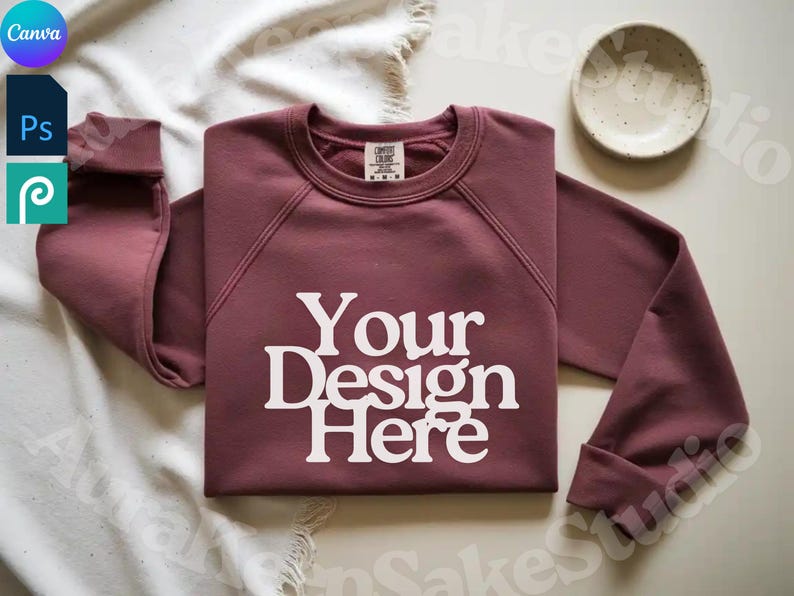 Pode incluir: Uma camisola de gola redonda bord&ocirc; com o texto "Your Design Here" em branco. A camisola &eacute; exibida em uma superf&iacute;cie branca com uma pequena tigela branca. As mangas est&atilde;o dobradas. Os logotipos Canva, Photoshop e Procreate est&atilde;o no canto superior esquerdo.