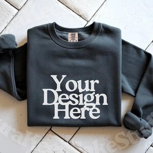 Pode incluir: Camisola cinza escuro com o texto "Your Design Here" em branco. A camisola de gola redonda de manga comprida &eacute; exibida em uma superf&iacute;cie de azulejos brancos. A camisa tem uma etiqueta na parte de tr&aacute;s do pesco&ccedil;o.
