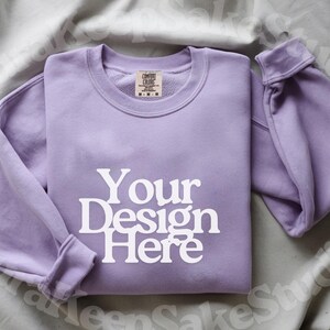 Pode incluir: Uma camisola de gola redonda lavanda com o texto "Your Design Here" em branco. As mangas est&atilde;o dobradas e a pe&ccedil;a de roupa est&aacute; cuidadosamente dobrada. Uma etiqueta &eacute; vis&iacute;vel na gola interior. A camisola &eacute; exibida numa superf&iacute;cie cinza claro.