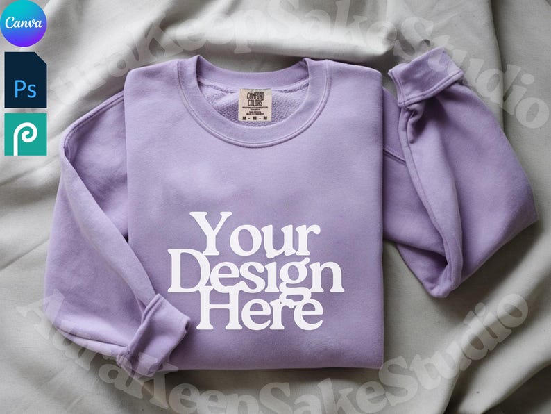 Pode incluir: Uma camisola de gola redonda lavanda com o texto "Your Design Here" em branco. As mangas est&atilde;o dobradas e a pe&ccedil;a de roupa est&aacute; cuidadosamente dobrada. Uma etiqueta &eacute; vis&iacute;vel na gola interior. A camisola &eacute; exibida numa superf&iacute;cie cinza claro.