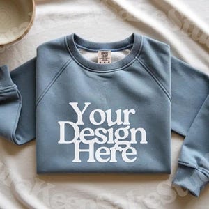 Pode incluir: Camisola azul clara com o texto branco "Your Design Here". Possui gola redonda e punhos canelados. Logotipos de software de design tamb&eacute;m s&atilde;o vis&iacute;veis.