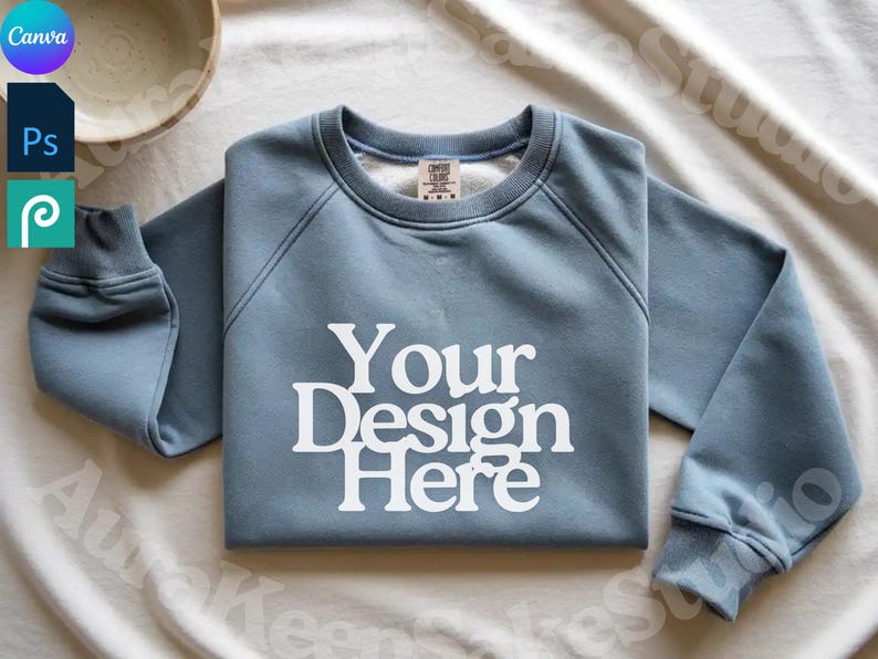 Pode incluir: Camisola azul clara com o texto branco "Your Design Here". Possui gola redonda e punhos canelados. Logotipos de software de design tamb&eacute;m s&atilde;o vis&iacute;veis.