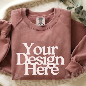 Pode incluir: Um moletom rosa empoeirado com o texto "Your Design Here" em letras brancas. O moletom tem gola redonda e mangas compridas. A etiqueta Comfort Colors &eacute; vis&iacute;vel.