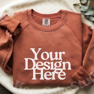 Pode incluir: Uma camisola cor de ferrugem com o texto "Your Design Here" em branco. A camisa de manga comprida est&aacute; dobrada e tem uma etiqueta na gola. A imagem &eacute; uma maquete de produto, provavelmente para um design de roupa.