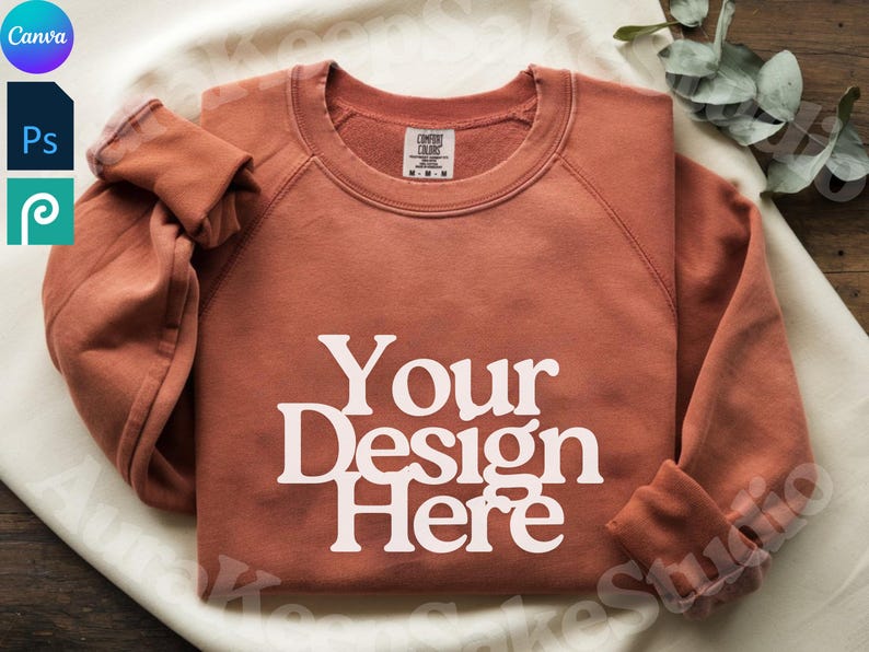 Pode incluir: Uma camisola cor de ferrugem com o texto "Your Design Here" em branco. A camisa de manga comprida est&aacute; dobrada e tem uma etiqueta na gola. A imagem &eacute; uma maquete de produto, provavelmente para um design de roupa.