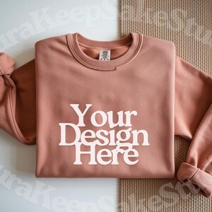 Pode incluir: Um moletom rosa empoeirado com o texto "Your Design Here" em letras brancas. O moletom tem gola redonda e mangas com punhos. O fundo &eacute; de cor neutra.