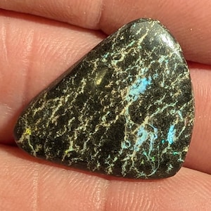 Könnte beinhalten: Ein polierter, dreieckiger schwarzer Opal-Edelstein mit einem einzigartigen Muster aus weißen Adern und schillernden blauen und grünen Lichtreflexen. Der Stein wird in einer Hand gehalten und zeigt seine natürliche Schönheit.
