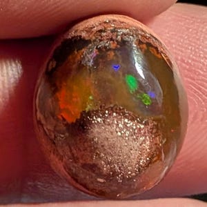 May include: A polished, oval-shaped opal gemstone. The stone displays a mix of colors, including orange, green, and blue, with a dark brown base and a textured, speckled surface. The opal has a glossy, reflective appearance.