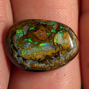 May include: An oval-shaped boulder opal gemstone featuring a dark brown ironstone matrix with vibrant flashes of iridescent green, blue, and gold color play across its polished, smooth, and slightly domed surface, showcasing natural mineral patterns.