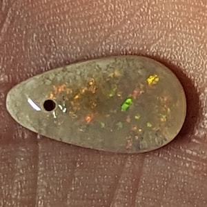 May include: A teardrop-shaped opal gemstone with a hole drilled through it. The opal has a milky white base with flashes of green, orange, and yellow.