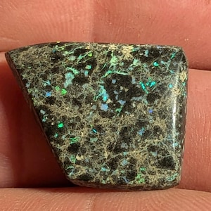 May include: A polished, irregularly shaped gemstone with a dark, speckled base. The stone features vibrant flashes of green, blue, and teal throughout its surface. The overall appearance is a mix of dark and iridescent colors.